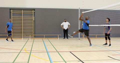 Diverse male volleyball team jumping and reaching at indoor court with coach watching