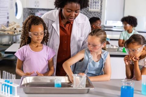 Stem teacher guiding elementary girls through hands-on science experiment with blue liquid