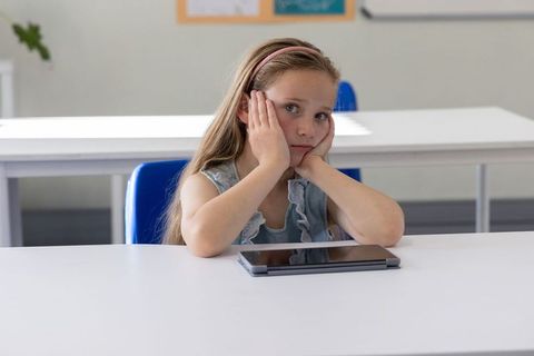 Bored schoolgirl resting face in hands at primary classroom desk with tablet and headband