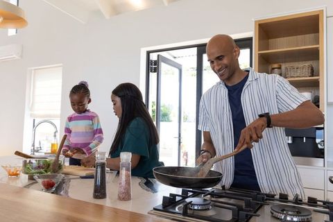 Sunlit modern kitchen family cooking together: father stirring pan, girls preparing salad