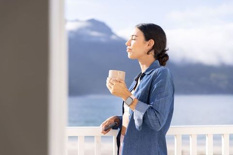 Woman enjoying morning coffee on balcony overlooking calm fjord wearing smartwatch