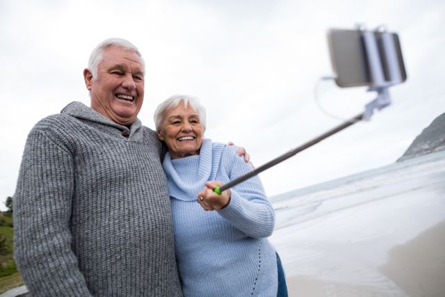 Senior couple enjoying a day at the beach, taking a selfie with a selfie stick. They are smiling and appear happy, wearing casual sweaters. Ideal for use in advertisements, travel brochures, retirement planning materials, and articles about senior living, technology use among elderly, and family bonding.