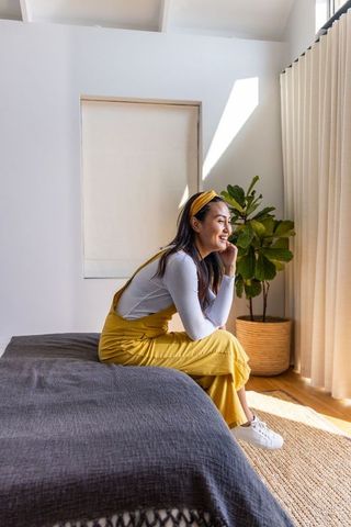 Smiling asian woman sitting on bed in sunlit minimalist bedroom wearing mustard overalls
