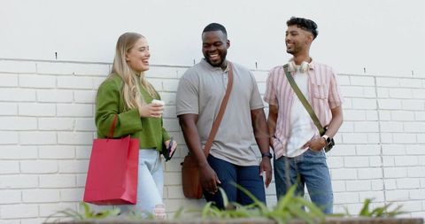 Diverse friends laughing and chatting while leaning on white brick wall with shopping bag
