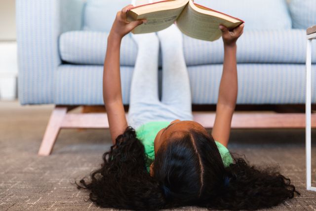 African american elementary schoolgirl reading book while lying on carpet in school play room. unaltered, childhood, education, reading, relaxation and school concept.