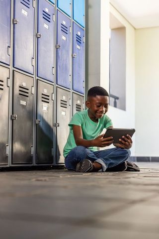 African american boy sitting in school hallway using tablet near lockers, smiling