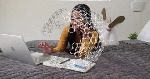 Remote worker calling and typing on laptop while lying on bed with hexagonal overlay