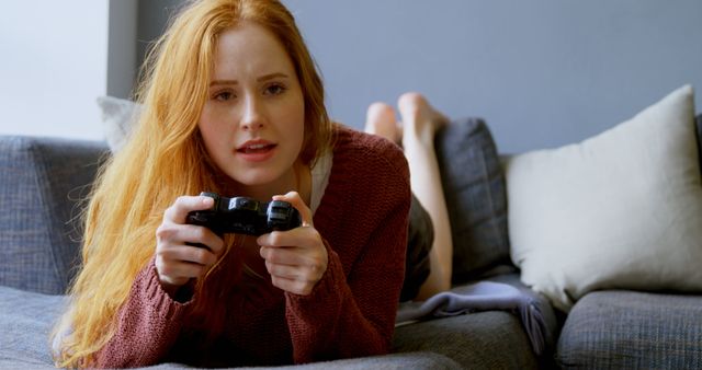 Young Caucasian woman enjoys gaming on the couch at home. She's fully engaged with a video game controller in her hands.