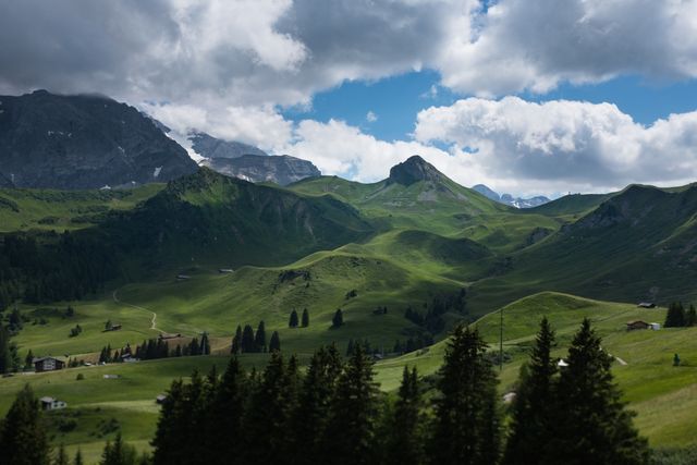Scenic view of an Alpine valley featuring lush green hills and mountains beneath a partly cloudy blue sky. Ideal for use in travel, tourism, nature, or adventure-related content, as well as for backgrounds and wallpapers.