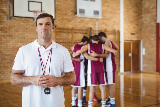 Confident basketball coach standing in gym with team huddling in background. Ideal for use in sports training materials, coaching guides, team-building articles, and athletic leadership promotions.