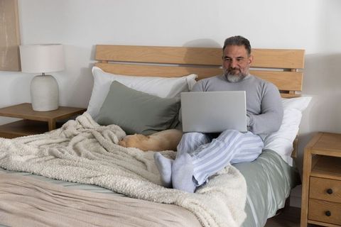 Mature man working from bed at home with laptop and sleeping dog in cozy neutral bedroom