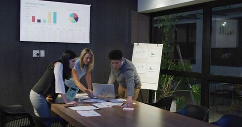 Collaborating team reviewing sales charts and laptop around conference table in modern office