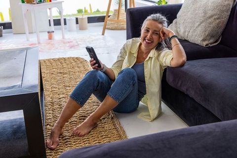 Senior woman relaxing on jute rug holding smartphone and smiling in modern living room