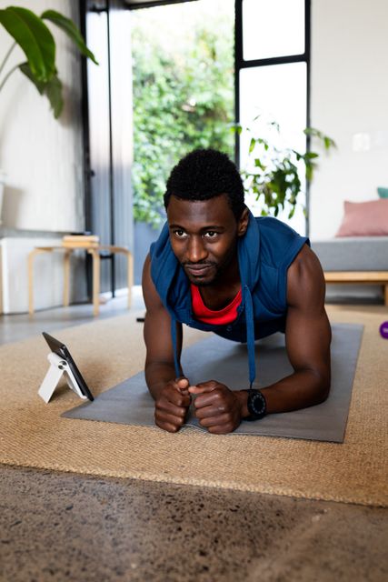 Vertical image of smiling african american man using tablet and exercising at home, copy space. Domestic life, living alone, communication, inclusivity and healthy lifestyle.