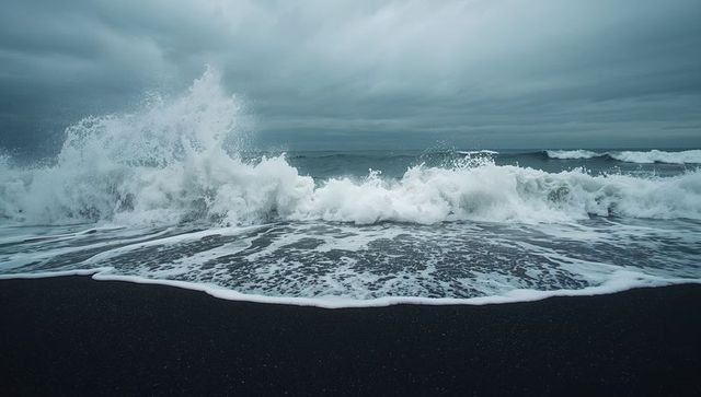 Crashing waves surging onto dark sand beach under stormy clouds with white seafoam spray