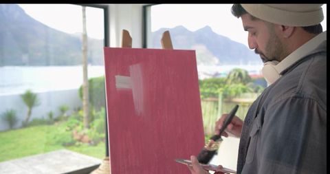Indian artist painting on canvas by window overlooking mountains and garden