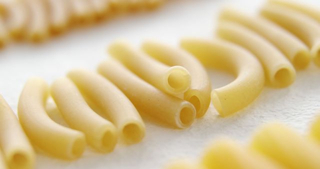 Uncooked macaroni pasta is arranged on a white surface, showcasing its tubular shape and smooth texture. Macaroni is a staple in Italian cuisine, often used in a variety of dishes from classic mac and cheese to hearty pasta salads.