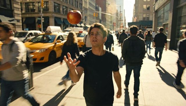 Tossing and catching red apple on sunlit downtown sidewalk with yellow taxis and crowd