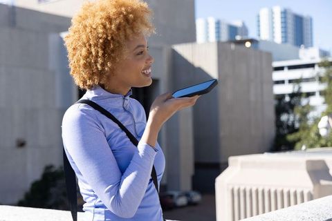 Young african american woman using smartphone voice assistant on rooftop with urban skyline