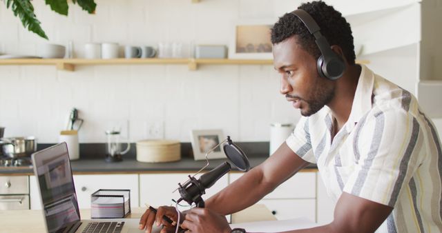 An African American man is recording a podcast at home. He is using a laptop and wearing headphones while speaking into a microphone. Modern kitchen environment in background with shelves and decor. Useful for illustrating remote work, podcasting, content creation, and technology in media production.