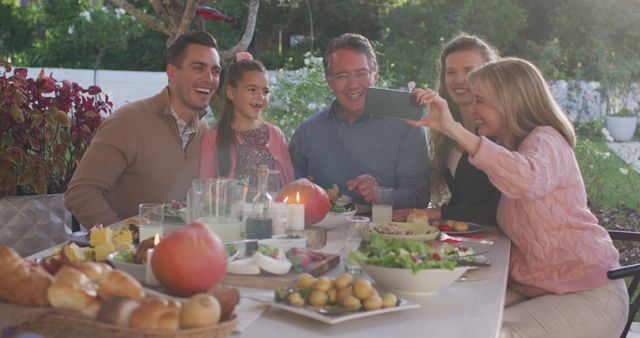 Family members enjoying dinner outdoors in their garden smiling for a selfie. Perfect for advertisements related to family bonding, summer activities, outdoor dining, celebrations, or promoting a healthy, happy lifestyle.