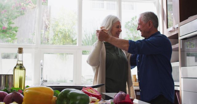 Senior couple enjoy dancing together in their bright, modern kitchen. Surrounded by fresh vegetables and cooking ingredients, they share a joyful moment. Ideal for use in campaigns related to healthy living, retirement planning, and promoting positive lifestyles during senior years.