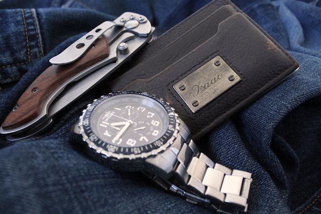 Depiction of men's accessory essentials including a high-end wristwatch with metallic band, a leather wallet with a name tag, and a sophisticated pocket knife with a wooden handle. Suitable for illustrating modern men's fashion, lifestyle blogs, and articles on everyday carry items. Can be used in advertisements for luxury accessories or promotional content for men’s fashion brands.