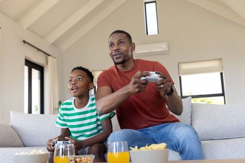 African american father and son playing video game on sofa with snacks and natural light