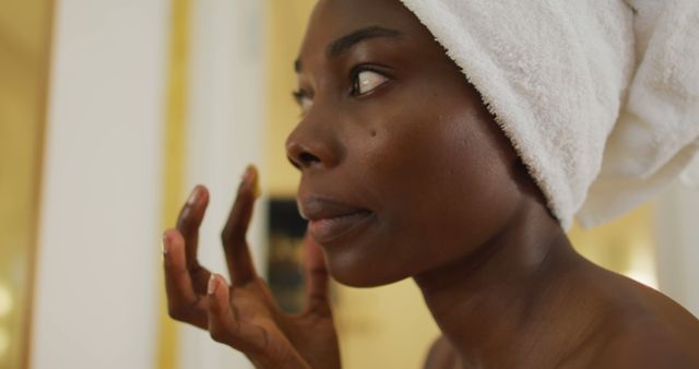 African american woman with towel watching in mirror and using cream on her face in bathroom - Download Free Stock Photos Pikwizard.com