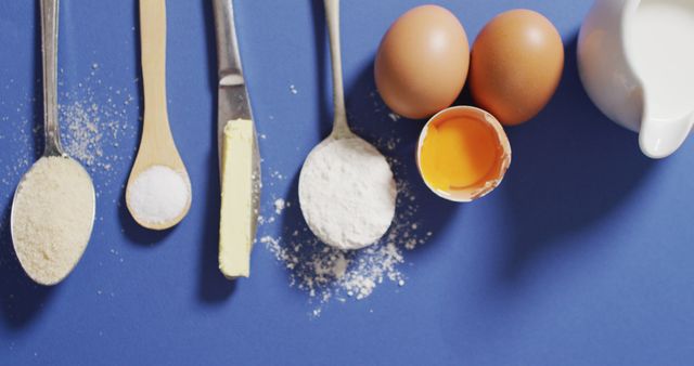 This image shows a flat lay of various baking ingredients, including two brown eggs, a halved egg with the yolk visible, flour, sugar, butter, and milk arranged neatly on a blue background. Ideal for use in cooking blogs, recipe cards, cooking tutorials, kitchen decoration, or culinary magazines. The neat arrangement provides a clean and organized look suitable for educational or marketing materials in the culinary field.