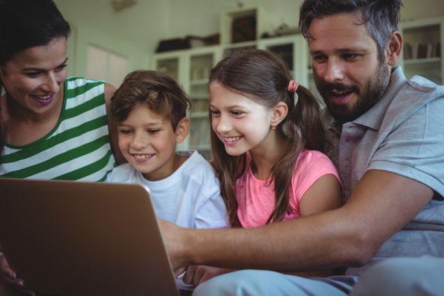 This image shows a happy family sitting together on a sofa in their living room, using a laptop. The parents and children are smiling and appear to be enjoying their time together. This image can be used for promoting family bonding, digital learning, home lifestyle, and technology use in family settings.