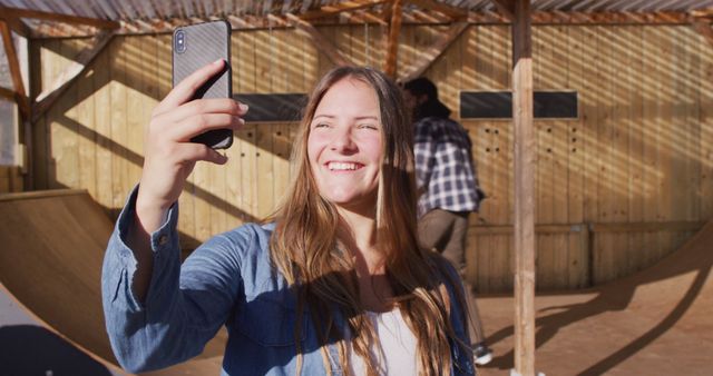 A young woman with long brunette hair is taking a selfie with her smartphone in an outdoor skatepark. She is smiling and appears happy, enjoying a sunny day. The surroundings include wood and corrugated metal structures. This stock photo can be used for projects related to social media, outdoor activities, modern lifestyle, and youth culture.