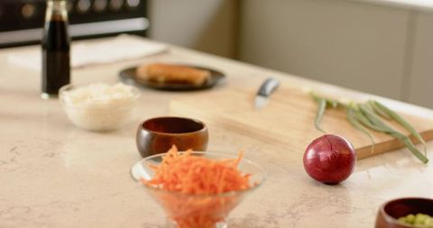 Preparing shredded carrot in glass bowl on quartz countertop with red onion and cutting board