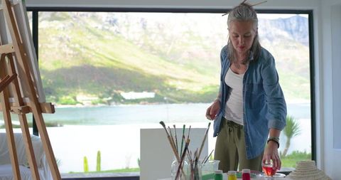 Mature woman arranging brushes in sunlit lakeside studio, painting supplies on table