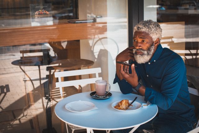 Senior African American man sitting at an outdoor cafe table, enjoying a cup of coffee and using his smartphone. He appears to be engaged in a conversation or recording a message. The scene suggests a modern, independent lifestyle, perfect for illustrating themes of technology use among seniors, urban living, and digital nomadism. Ideal for use in articles or advertisements related to senior lifestyle, technology, and urban culture.