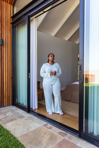 African american woman standing in sliding door holding mug in sunlit bedroom retreat