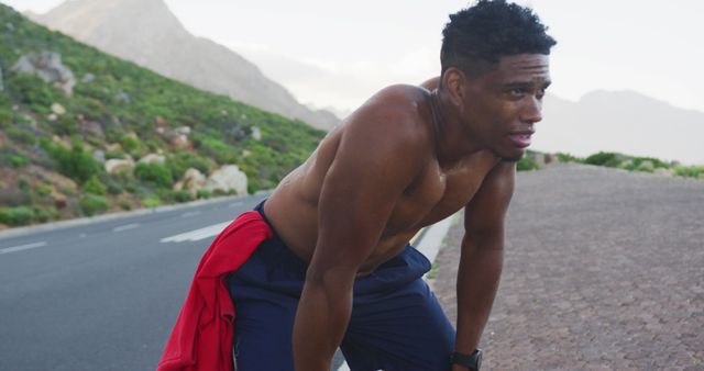 African American man pausing to catch his breath on a mountain road after an intense run. Appears sweaty and worn out, showing hard work and perseverance. This can be used for promoting fitness, athletic brands, motivation, or outdoor activity promotions.