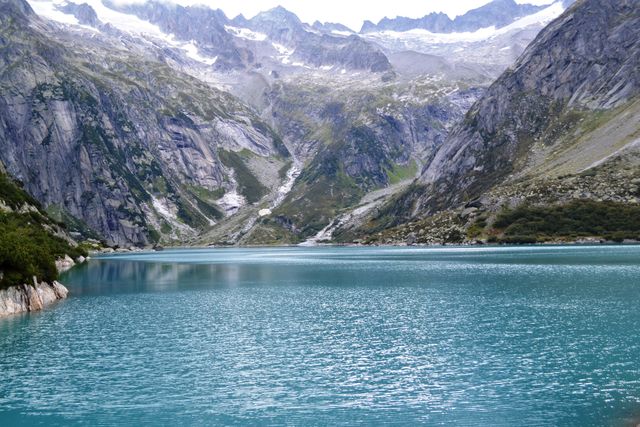 Alpine lake with clear blue water surrounded by rocky hills and snow-covered mountain peaks in the background. Perfect for use in travel brochures, nature websites, outdoor adventure blogs, and wallpapers depicting natural beauty and tranquility of wilderness.
