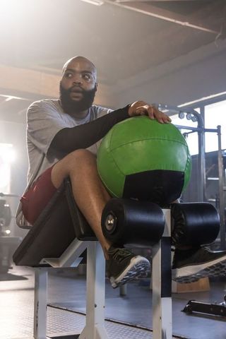 African american man sitting on decline bench holding green medicine ball, focused athlete