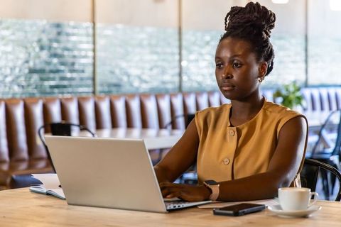 African woman working on laptop in cafe, focused professional productivity and notetaking