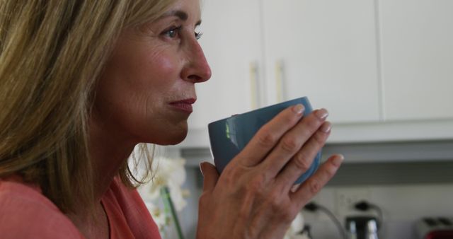 A mature woman sips a hot beverage, enjoying a moment of relaxation in her home kitchen. Image great for use in lifestyle blogs, advertisements for home products, wellness articles, and retirement planning materials.