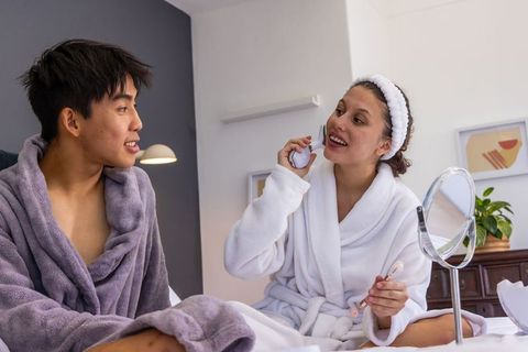 Couple enjoying cozy morning skincare routine in plush robes on bed with mirror and facial device
