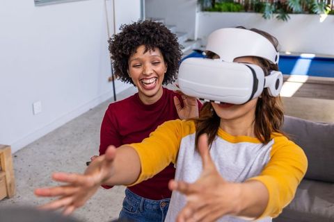 African american woman laughing while hispanic friend guiding vr headset user at home