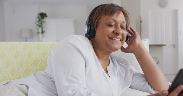 Senior woman enjoying leisure time at home, listening to music through headphones, smiling. Ideal for promoting products related to relaxation, retirement living, senior lifestyle, home comfort, and personal audio devices.
