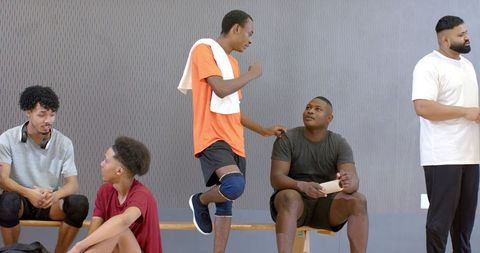 Multiracial male teammates relaxing and chatting on bench in indoor gym after practice