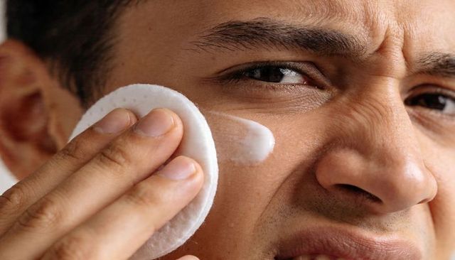 Hispanic man pressing cotton pad with cleanser on cheek closeup showing skincare texture