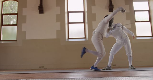 Two athletes are engaging in a fencing match in a well-lit gymnasium. They are wearing traditional white fencing uniforms and protective gear. One fencer lunges towards the other, demonstrating skill and agility. This can be used for topics related to sportsmanship, physical training, martial arts, competition, and team activities. Ideal for promoting fencing clubs, sports events, or fitness programs.