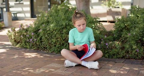 Young girl sitting cross-legged in courtyard reading and flipping red notebook pages