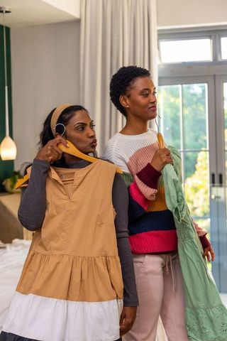 Indian and african american friends comparing dresses in cozy bedroom, choosing outfits