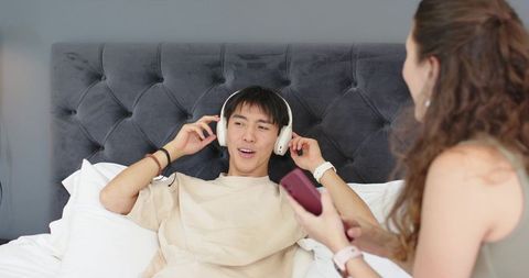 Young man lounging on bed listening to headphones while partner holding smartphone